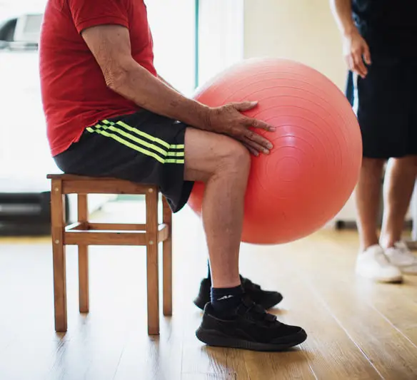 Paziente anziano che esegue esercizi di mobilità e coordinazione seduto su uno sgabello con l'ausilio di una fitball rossa.
